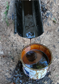 Bilden visar en stråle av mörk tjära som rinner från en ränna ner i en plåtburk