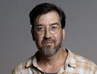 A man with dark hair, beard and moustache, and glasses look into the camera.