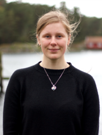 Tjej med svart tröja och silverhalsband står framför havet, en brygga och ett rött hus 