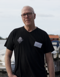 Man wearing a black t-shirt and a name tag stands in front of a pier with boats