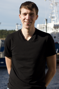 Ung kille med svart t-shirt står framför ett fartyg i en hamn 