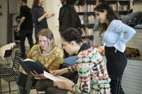 En tjej i gul tröja läser, en annan tjej med uppsatt hår sitter bredvid med ett papper i handen.