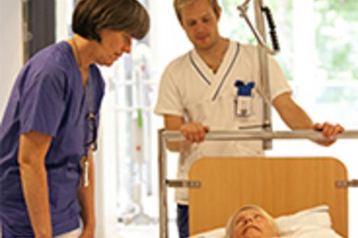 Två personer, sjukvårdspersonal, står vid patient i sjukhussäng