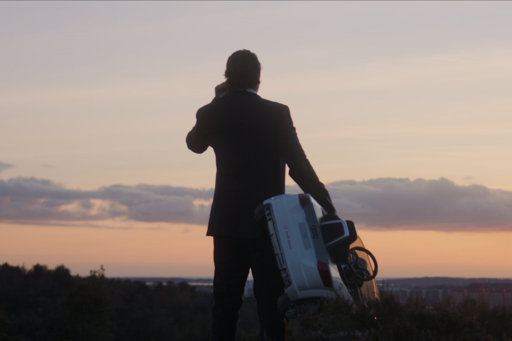A man is holding a large toy car and looking out over the evening sky where a drone is flying
