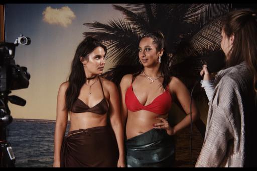 Två lättklädda kvinnor står framför en filmkamera och en tropisk kuliss