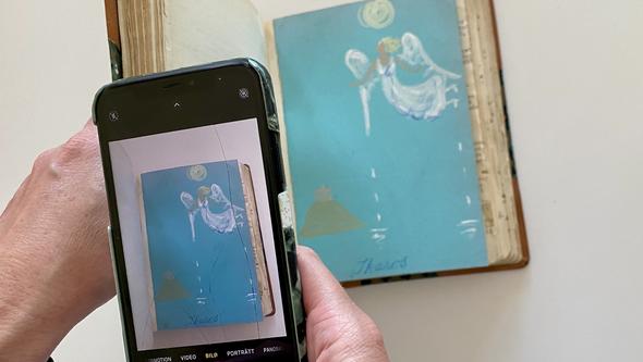 En tecknng av vingar (Ikarosillustration) i en bok. Fotograferas av mobilkamera som betrataren ser igenom
