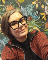 Porträtt av Charlott Sellberg, leende med glasögon och mot mönstrad tapet