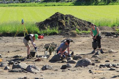 An excavation site with three people digging, in the background a green field, photo. 